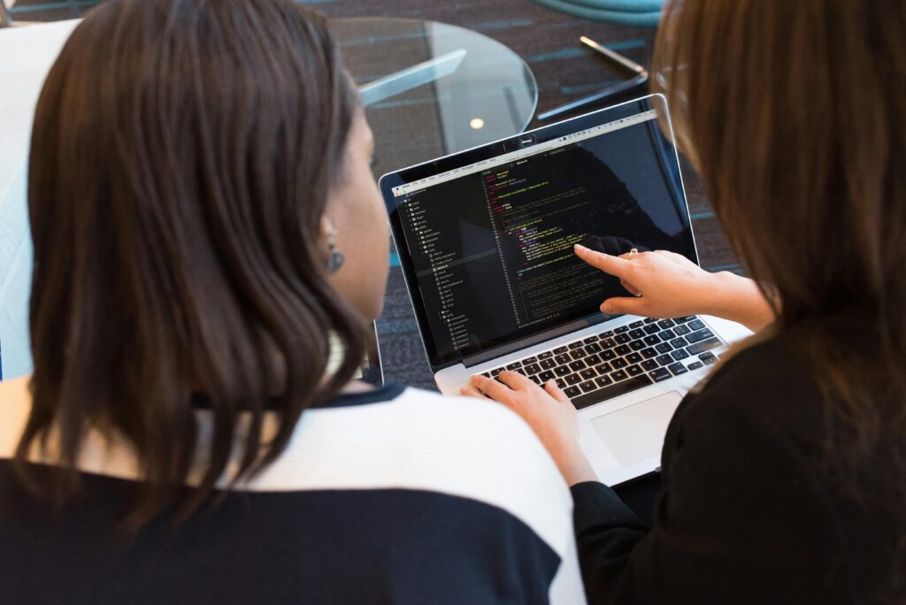 A stock image showing: Two woman sitting in front of a laptop that displays code. One woman is pointing at the code and explaining it to the other woman.