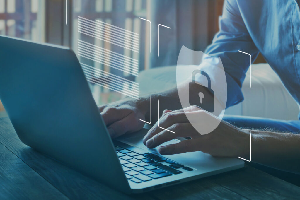 a stock image showing: A man sitting in front of a laptop with his hands on the keyboard. Above the keyboard a lock inside a shield is visible.